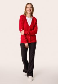 Cardigan rosso con scollo a V e chiusura con bottoni, abbinato a pantaloni neri a zampa d'elefante e sneakers bianche, su uno sfondo neutro.