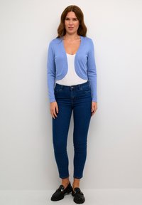 Ljusblå kortärmad cardigan över en vit topp, ihop med mörkblå skinny jeans och svarta loafers. Slät stickad textur, enkel design.