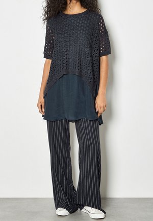 Femme portant un haut court en crochet noir sur une tunique sombre, un pantalon large à fines rayures noires et blanches, et des baskets blanches, debout contre un mur uni.