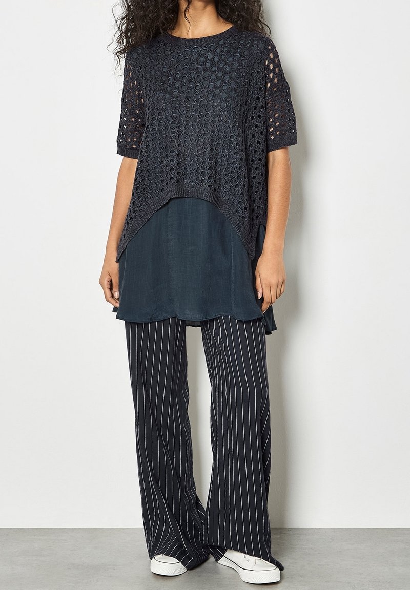 Femme portant un haut court en crochet noir sur une tunique sombre, un pantalon large à fines rayures noires et blanches, et des baskets blanches, debout contre un mur uni.