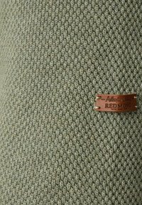 Strickstoff in gedämpftem Grünton mit strukturiertem Muster; zeigt ein braunes Lederetikett mit eingeprägtem Text "Authentic Clothing REDMOND."