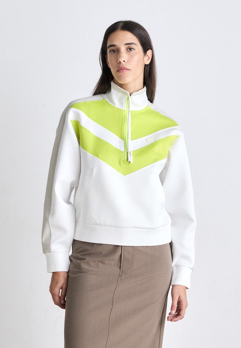 Weißer Sweatshirt mit hohem Kragen, der auffällige limonengrüne und weiße Zickzackmuster aufweist. Der Stoff scheint weich und hat eine entspannte Passform.