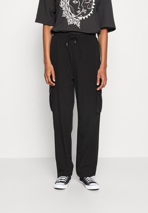 Person iført sorte cargobukser med sidelommer, sort T-shirt med hvid sol- og måne grafik, og sorte og hvide sneakers.