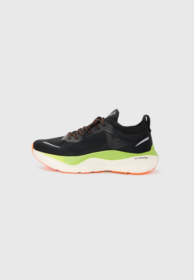 Black athletic sneaker with green and white sole, orange accents, textured sides, and laces on a plain light gray background.