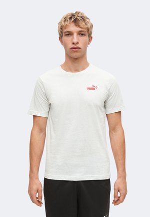 Jeune homme aux cheveux blonds courts, portant un t-shirt blanc Puma et un pantalon noir, debout devant un fond clair uni.