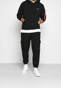 Felpa nera con tasca a marsupio e tasca con zip, abbinata a pantaloni cargo neri con due tasche laterali e sneakers bianche.