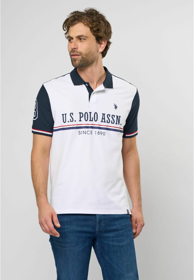Hvid polo-shirt med marineblåt krave og ærmer, prydet med et stort logo, røde og marineblå striber og korte ærmer. Bæres med blå jeans.