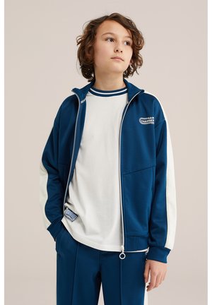 Adolescente con capelli ricci che indossa una giacca con zip blu e bianca e pantaloni abbinati, guarda leggermente verso l'alto e di lato.