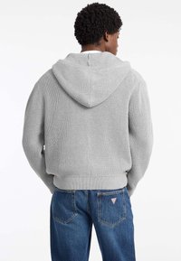Sudadera gris de punto con textura canalé, corte oversize y capucha amplia. Lleva puesta sobre unos vaqueros de mezclilla azules con un parche de logo en el bolsillo.