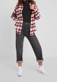 Camicia di flanella a quadretti rossa, bianca e blu, indossata sopra un dolcevita nero. Jeans scuri a vita alta con risvolti e scarpe da ginnastica bianche.
