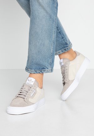 Persoon draagt beige Adidas-sneakers met witte zolen en blauwe spijkerbroek, staand op een lichtgrijze vloer tegen een effen achtergrond.