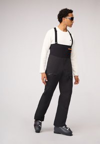 Schwarze Skihosen mit verstellbaren Trägern, einer Reißverschlusstasche und einer glatten Textur. Kombiniert mit grauen Skischuhen und einem cremefarbenen Langarmshirt.