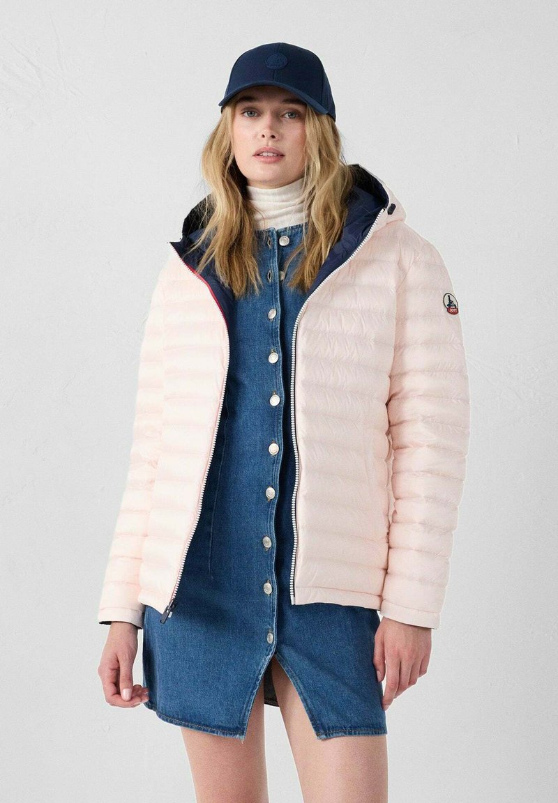 Giacca imbottita rosa chiaro con chiusura a zip, cappuccio foderato in blu navy, indossata sopra un vestito in denim con bottoni. Il cappello presenta un logo circolare.