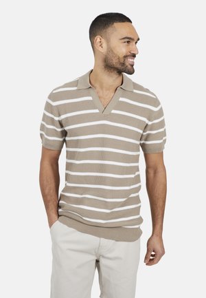 Homme avec une barbe courte portant un polo à manches courtes beige et blanc à rayures et un pantalon clair, souriant et regardant vers la droite.