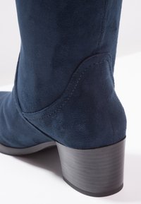 Gros plan sur une botte cheville en daim bleu marine avec un talon bloc empilé et un détail de couture incurvée à l'arrière.