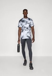 Man met een zwart-wit tie-dye shirt, zwarte Nike leggings en zwarte sneakers, die een zwarte Nike waterfles vasthoudt, staand op een witte vloer.