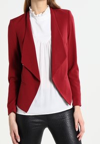 Blazer rosso con un design aperto sul davanti, tessuto morbido e vestibilità slim. Indossato sopra una camicetta bianca con scollo arruffato. Pantaloni neri in tessuto testurizzato.