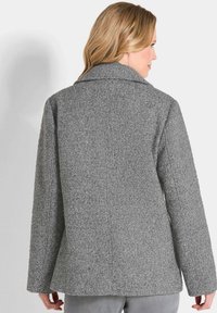 Frau, die ein graues, strukturiertes Wollblazer trägt, von hinten gezeigt, mit langen Ärmeln und einem Reverskragen.