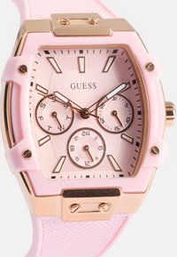 Reloj con correa de goma rosa, caja cuadrada, acentos en oro rosa, y una esfera texturizada que presenta tres subesferas y marcadores de horas en negrita.