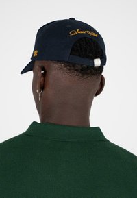 Marineblauwe pet met gele geborduurde tekst "Social Club," verstelbare band en metalen hardware. Draag het met een groene gestructureerde trui.