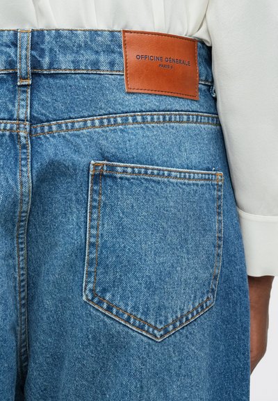 Jeanși de denim de culoare albastru deschis cu un etichetă din piele "OFFICINE GÉNÉRALE PARIS 6" pe spate, având cusături portocalii și un singur buzunar la spate.