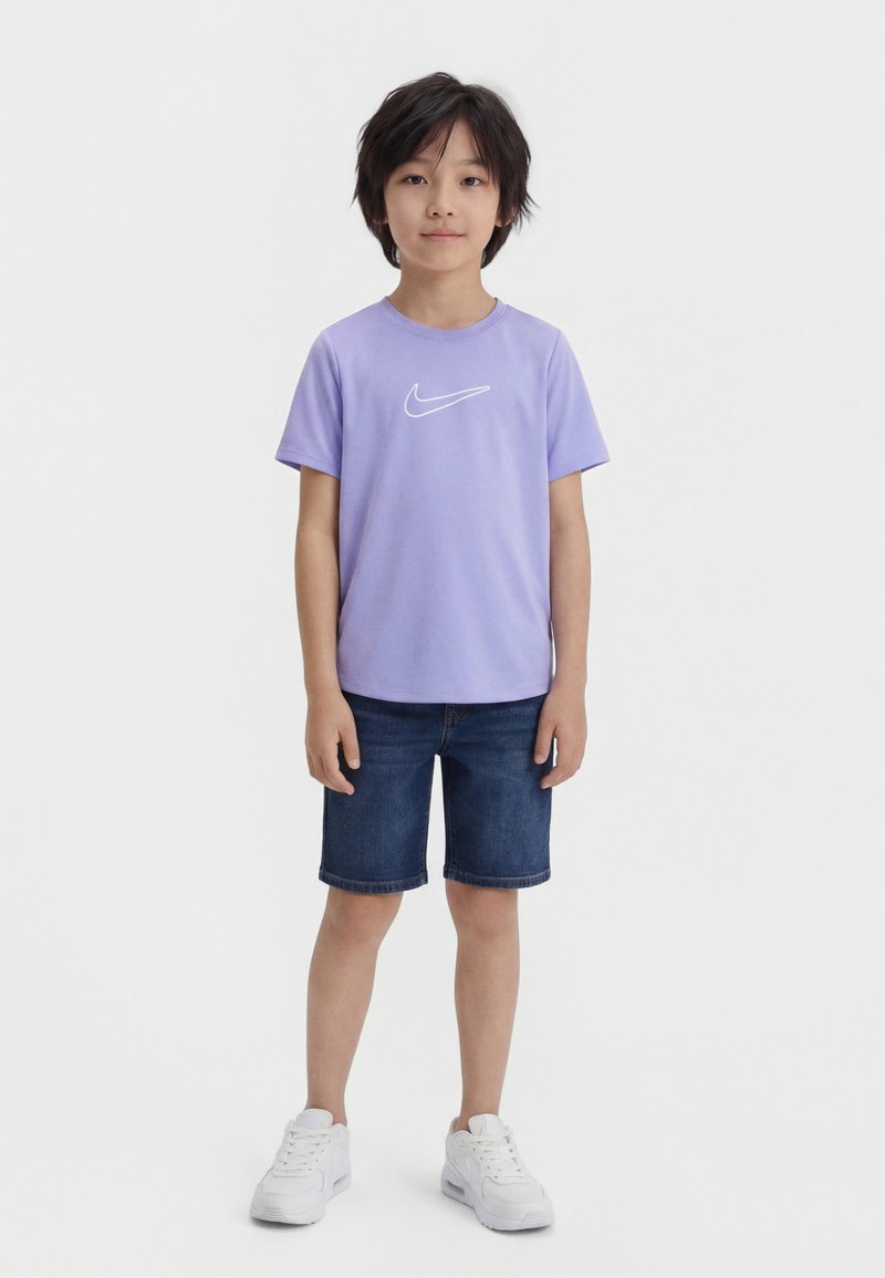 Αγόρι όρθιο, φοράει ανοιχτό μοβ μπλουζάκι Nike, σκούρες μπλε τζιν σόρτς και λευκά αθλητικά παπούτσια, σε λευκό φόντο.