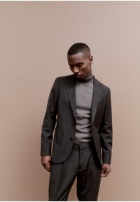 Grijze pinstripe kostuum met een slim fit, gecombineerd met een lichtgrijze coltrui. De stof lijkt glad, op maat gemaakt en gestructureerd. Neutrale achtergrond.