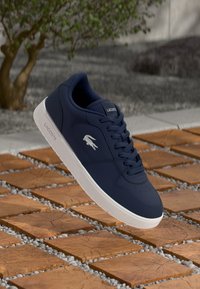 Marineblaue Leder sneaker mit weißer Gummisohle. Merkmale sind eine perforierte Zehenkappe, runde Schnürsenkel und das Lacoste-Logo an der Seite.