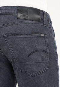 Mörkgrå denimjeans med en läderremmärkning som har "RAW". Distinkt sömnad på bakfickorna och en klassisk femficksdesign.
