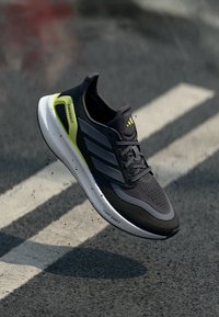 Sort atletiksko med grå accenter og en lys gul hæl, der har en tekstureret mesh-overdel og en hvid mellemsål mærket "LIGHT BOOST."