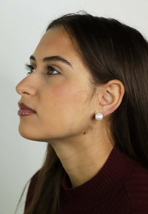 Jeune femme aux longs cheveux noirs, portant une boucle d'oreille en perle et un col roulé côtelé bordeaux, regardant vers la gauche sur un fond uni.