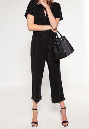 Femme portant une combinaison noire et des talons à bride à la cheville, tenant un sac à main noir avec un bras croisé sur sa taille.