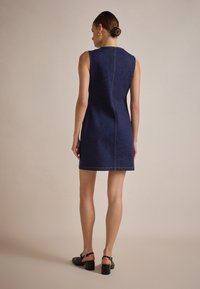 Robe en denim bleu foncé sans manches. Présente des coutures visibles et une silhouette ajustée. Le modèle porte des sandales à talons noires.