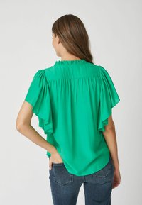 Zelená blúzka s krátkymi volánovými rukávy, gathered výstrihom a odvádzajúcim dizajnom. Mäkko, ľahký materiál s hladkou textúrou.