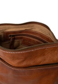 Interno di una borsa in pelle marrone con una fodera beige, scomparti con zip e cuciture bianche. Presenta una tasca piccola e un'etichetta.