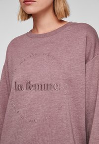 Bordeaux-coloured huppari, jossa on kohokuvioidut sanat "la femme" keskellä, ympäröitynä sanoilla "ihana", "pehmeä" ja "naisellinen", rento malli.