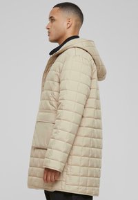 Beige quiltet dunjakke med hætte, vandret syninger, frontlomme og lange ærmer. Glat materiale med en blank finish.