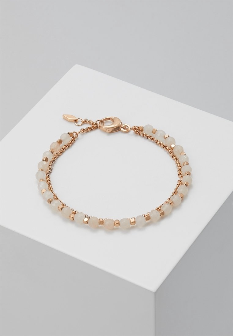 Délicat bracelet chaîne en or rose avec petites perles blanches rondes et fermoir mousqueton reposant sur une surface géométrique blanche.