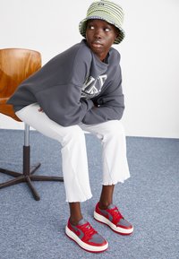 Baskets rouges et grises avec semelles blanches, pantalons blancs côtelés et un sweat-shirt gris. Un chapeau seau rayé vert et blanc complète le look.