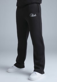 Pantalon de survêtement noir avec poches latérales, arborant un logo brodé blanc. Matière douce, coupe droite et taille côtelée.