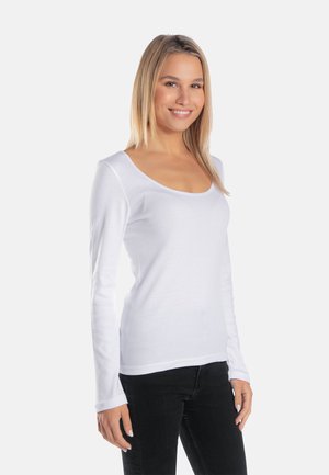 Młoda kobieta uśmiechająca się, ubrana w biały, długi rękaw, bluzkę z okrągłym dekoltem oraz czarne spodnie, stojąca na tle jednolitego jasnego tła.