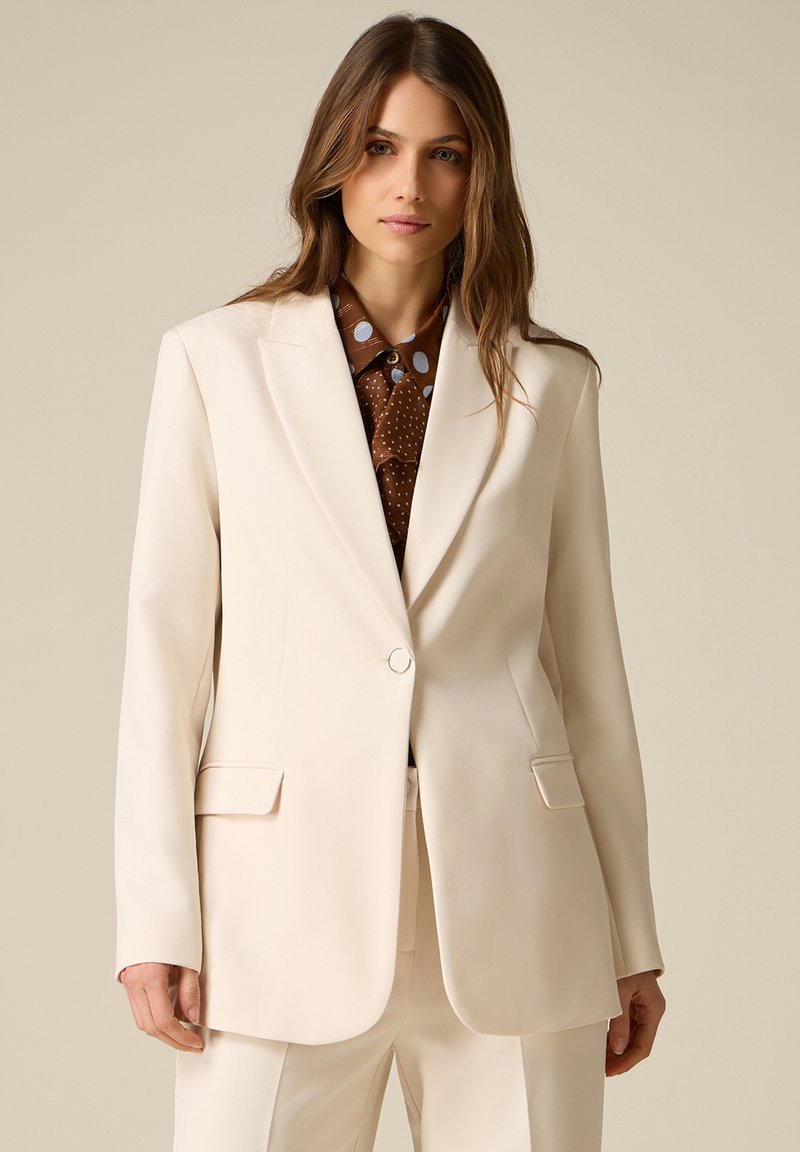 Mujer vestida con un traje crema y una chaqueta de un solo botón sobre una camisa marrón con lunares, de pie frente a un fondo neutro beige.