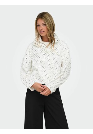 Femme aux cheveux blonds ondulés portant une veste blanche à pois et un pantalon noir à jambes larges, debout devant un fond uni.
