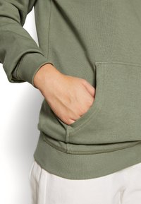 Hand in de kangoeroezak van een olijfgroene sweatshirt met geribbelde manchetten en zoom, gecombineerd met witte broek.