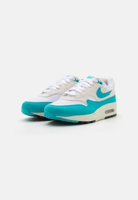 Tênis Nike Air Max 1 em branco e turquesa. Possui parte superior em malha, detalhes em camurça, unidade de ar visível e sola de borracha preta.