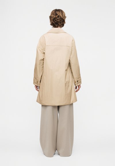 Άτομο με κοντά σγουρά μαλλιά που φοράει μακρύ μπεζ παλτό και πλατύ παντελόνι ανοιχτού γκρι χρώματος, στέκεται στραμμένο προς ένα λευκό φόντο.