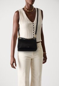 Persona che indossa un top senza maniche a maglia cable color crema e pantaloni con collana d'oro e borsa a tracolla nera con dettagli dorati.