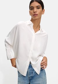 White oversized button-up shirt with long sleeves, made of smooth fabric. The shirt features a collar and subtle side slits at the hem.
