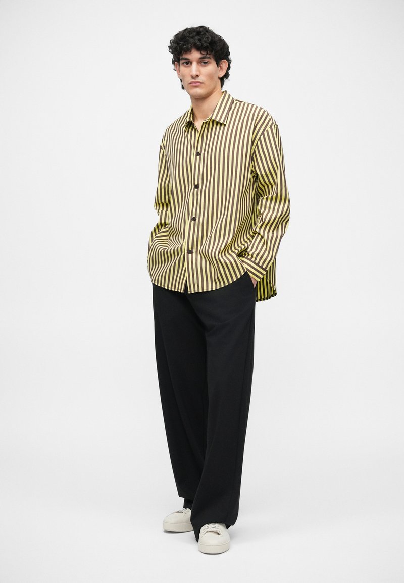 Striped shirt in yellow and brown, with a relaxed fit and button-down front. Paired with loose black trousers and white sneakers.