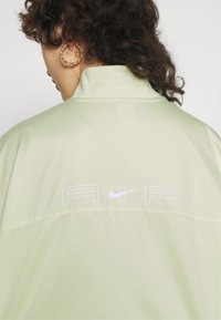 Ljusgrön jacka med hög krage. Har vit text "AIR" och Nike-logotyp på baksidan. Mjuk textur, avslappnad design.